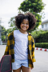 Smiling girl standing with skateboard on footpath - IKF00379
