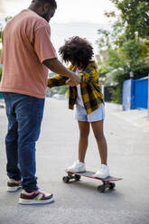 Father assisting daughter standing on skateboard at footpath - IKF00386