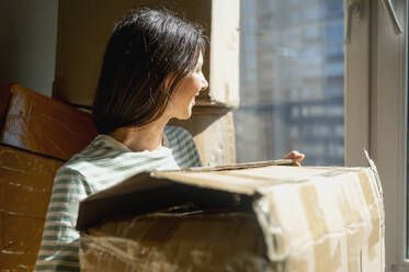 Lächelnde Frau schaut aus dem Fenster und hält einen Karton zu Hause - ANAF01304