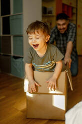 Playful father pushing son in cardboard box at home - ANAF01317