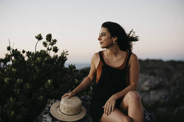 Smiling woman with hat sitting on rock - LHPF01578