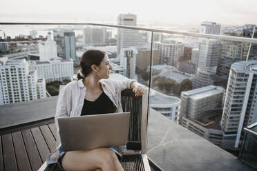 Freelancer with laptop sitting on chair on rooftop - LHPF01590