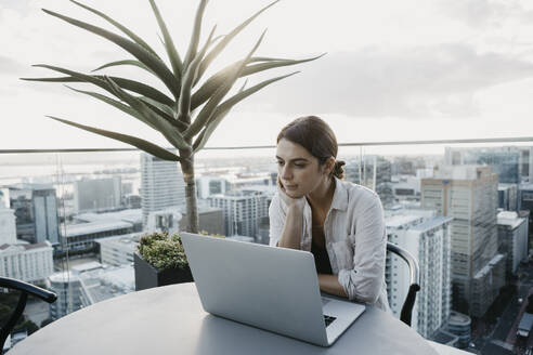 Woman with hand on chin sitting with laptop on rooftop - LHPF01595