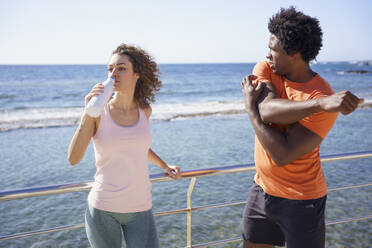 Man exercising with girlfriend drinking water by railing in coastal area - JSMF02743