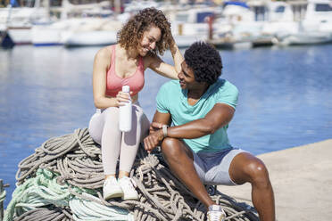 Mann zeigt seiner auf einem Seil im Hafen sitzenden Freundin eine intelligente Uhr - JSMF02749