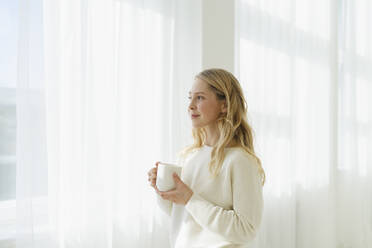 Smiling woman holding coffee cup by translucent curtain - SEAF01878