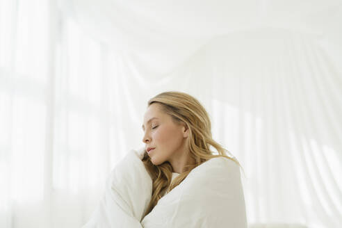 Frau mit geschlossenen Augen, eingewickelt in eine weiße Decke durch einen durchsichtigen Vorhang - SEAF01882