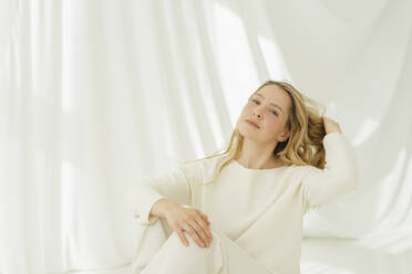 Blond woman with hand in hair sitting by translucent curtain - SEAF01894