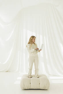 Blonde Frau mit Laptop auf einer Couch vor einem durchsichtigen Vorhang stehend - SEAF01900