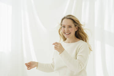 Lächelnde Frau mit blondem Haar tanzt vor einem weißen durchsichtigen Vorhang - SEAF01925