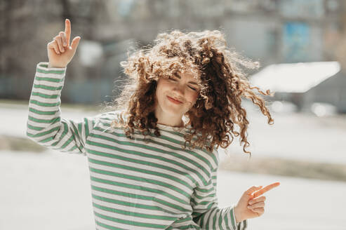 Smiling woman with curly hair dancing at sunny day - ANAF01330