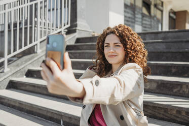 Lächelnde Frau nimmt Selfie durch Smartphone sitzen auf Stufen - ANAF01334