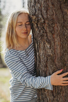 Junge Frau mit geschlossenen Augen umarmt Baum im Wald - ANAF01351