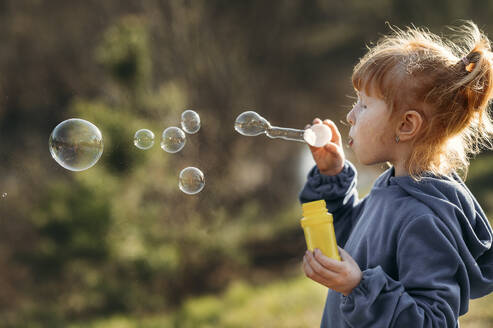 Girl with red hair blowing soap bubbles in forest on sunny day - ANAF01353