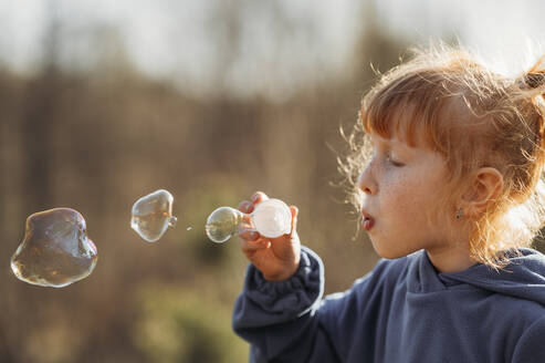 Girl with red hair blowing soap bubbles on sunny day - ANAF01354