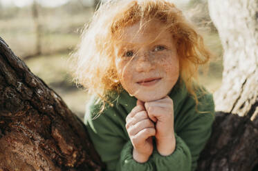 Smiling redhead girl with freckles leaning on tree - ANAF01363
