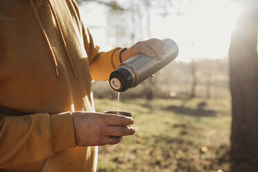 Mann gießt Tee aus isoliertem Getränkebehälter im Wald an einem sonnigen Tag - ANAF01367