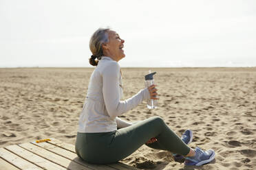 Frau mit Wasserflasche sitzend und lachend am Strand - EBSF03248