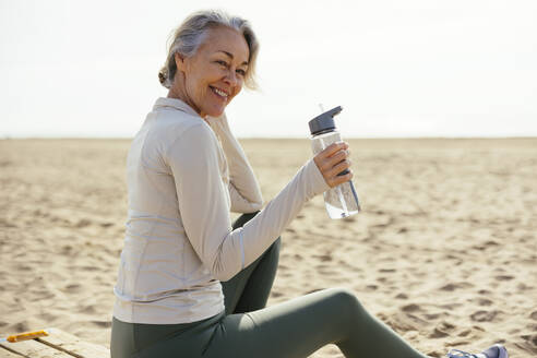 Smiling woman holding water bottle sitting at beach - EBSF03250