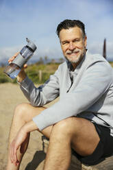 Lächelnder Mann mit Wasserflasche am Strand sitzend - EBSF03253