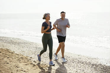 Lächelndes reifes Paar joggt zusammen in der Nähe des Ufers am Strand - EBSF03254