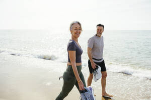 Lächelnde Frau geht mit Mann mit Sportschuhen am Strand spazieren - EBSF03269