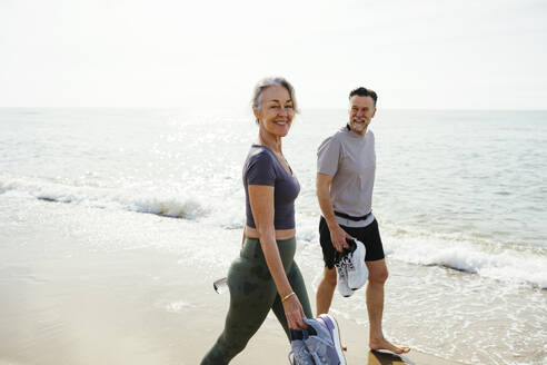 Smiling woman walking with man holding sports shoes at beach - EBSF03269