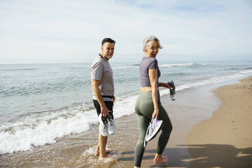 Mature couple holding sports shoes walking near shore at sunny day - EBSF03270