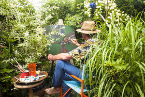 Man painting amidst plants sitting on chair in garden - HHF05883