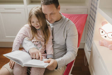 Girl sitting with father reading book at home - VIVF00769