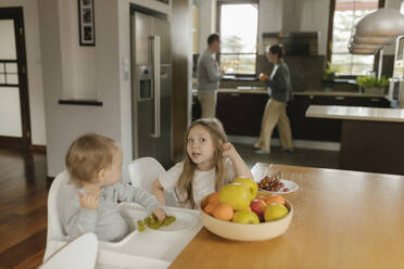 Siblings by fruit bowl at dining table with parents in background - VIVF00801