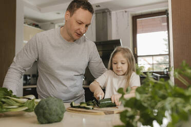 Father teaching daughter to cut zucchini in kitchen at home - VIVF00806