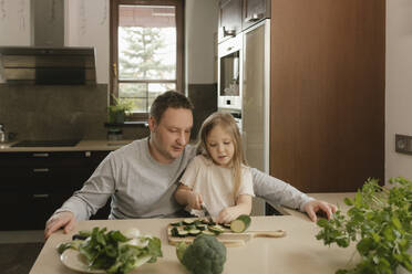 Father sitting with daughter cutting zucchini at kitchen table - VIVF00807