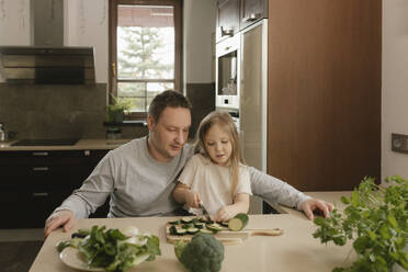 Vater sitzt mit Tochter beim Schneiden von Zucchini am Küchentisch - VIVF00807