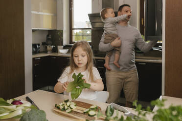 Mädchen hält Bok Choy mit Vater, der seinen Sohn im Hintergrund trägt, zu Hause - VIVF00814