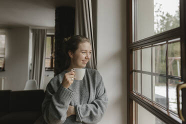 Smiling woman with coffee cup looking out of window at home - VIVF00815