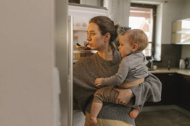 Mutter mit Baby in der Hand schaut in den Kühlschrank in der Küche zu Hause - VIVF00840
