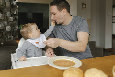 Vater füttert einen kleinen Jungen am Esstisch mit Suppe - VIVF00843