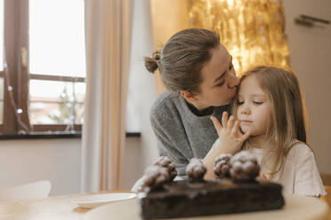 Mother kissing daughter by chocolate cake at table - VIVF00851