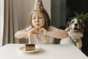 Mädchen macht Herz Geste und bläst Kerze auf Kuchen zu Hause - VIVF00862