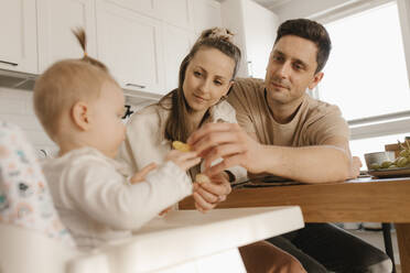 Father and mother feeding fruit to daughter sitting at home - VIVF00884