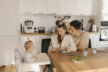 Mutter und Vater betrachten die süße Tochter, die zu Hause auf dem Hochstuhl Obst isst - VIVF00886