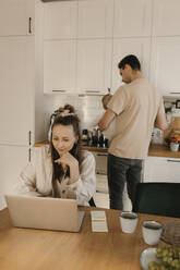 Woman working on laptop in kitchen with family in background at home - VIVF00888