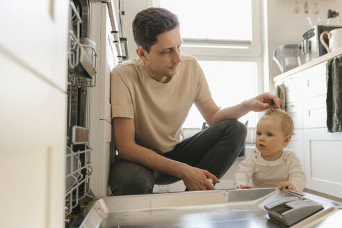 Cute baby girl and father loading utensils in dishwasher in kitchen at home - VIVF00892