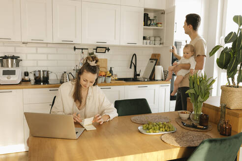 Eine Frau arbeitet an einem Laptop, während ein Mann im Hintergrund seine Tochter trägt und Hausarbeiten erledigt - VIVF00893