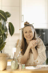 Lächelnde junge Frau, die einen Pinsel hält und sich zu Hause an einen Tisch lehnt - VIVF00910