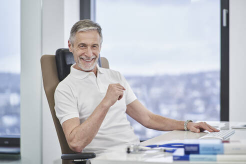 Portrait of smiling doctor sitting at desk in medical practice - RORF03463