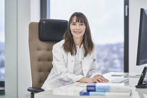 Porträt einer lächelnden Ärztin, die in einer Arztpraxis am Schreibtisch sitzt - RORF03482