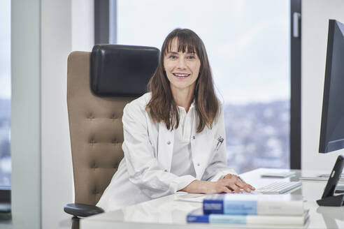 Portrait of smiling female doctor sitting at desk in medical practice - RORF03482