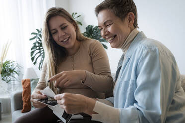 Happy pregnant woman sharing ultrasound photos with mother at home - AAZF00399